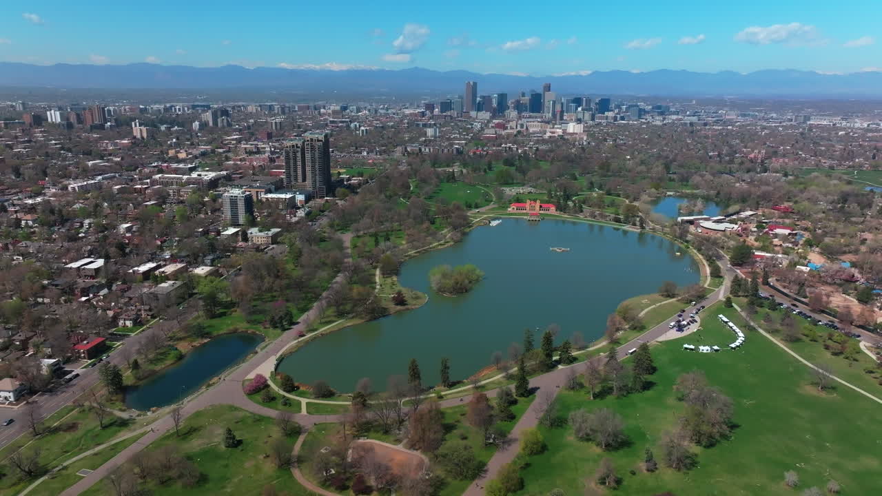 City Park Lodo Denver Colorado Lake Pavilion vibrant spring summer aerial drone sunny blue sky snow capped Rocky Mountains front range cityscape green lush grass trees blossom backwards pan up motion