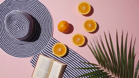 summer flat lay with beach hat, oranges, book, and palm leaf on pink background