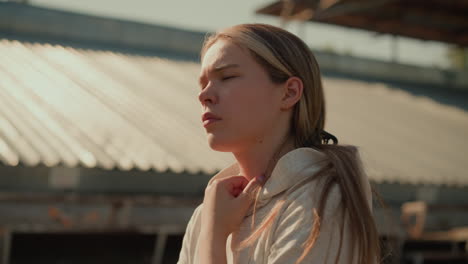 close-up of a thoughtful young woman walking outdoors in stadium, hand gently touching her hoodie, with a blurred background and soft sunlight