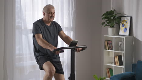 Entrenamiento-Cardiovascular-Matutino-En-Bicicleta-Estática-Un-Hombre-De-Mediana-Edad-Está-Haciendo-Deporte-En-Su-Sala-De-Estar-El-Fin-De-Semana-Sentado-En-Una-Bicicleta-Estática