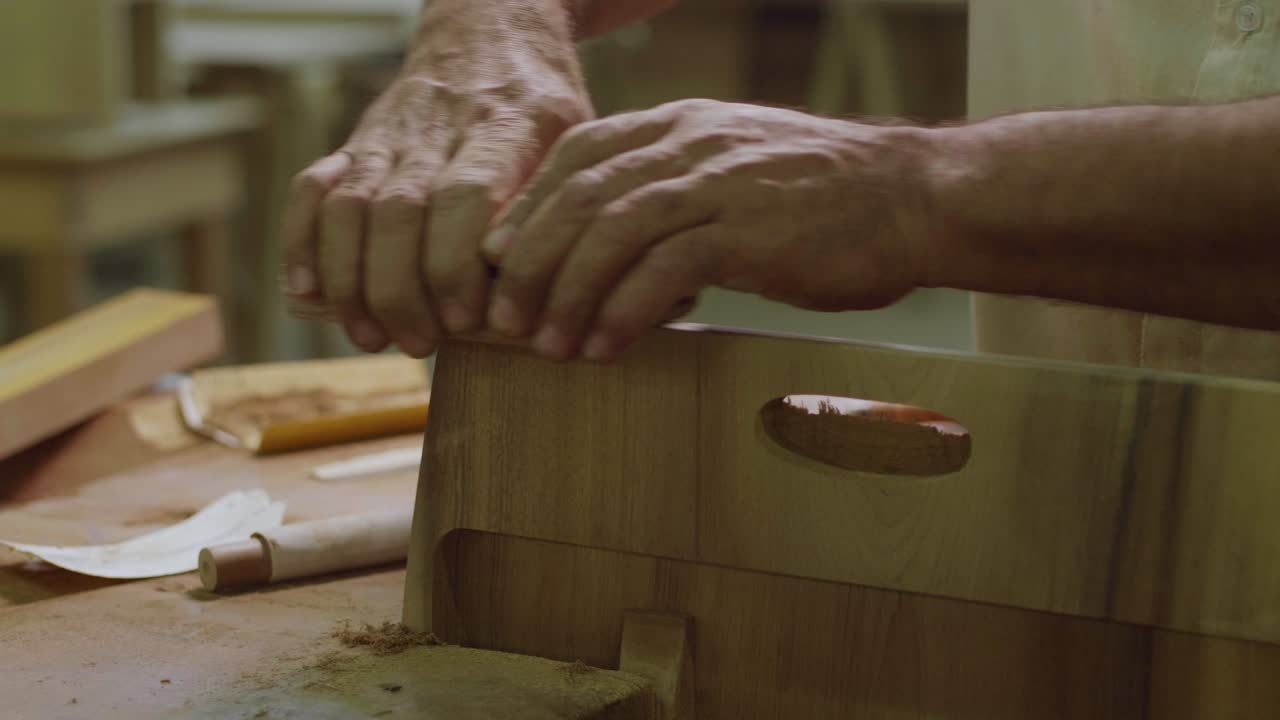 A close-up of hands working on a wooden piece with a carved handle, likely assembling or finishing a handcrafted furniture item on a workbench, surrounded by woodworking tools.