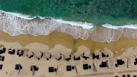 Top-down-shot-of-waves-crashing-in-over-golden-sand-beach-with-sun-beds-lining-the-edge-of-the-water