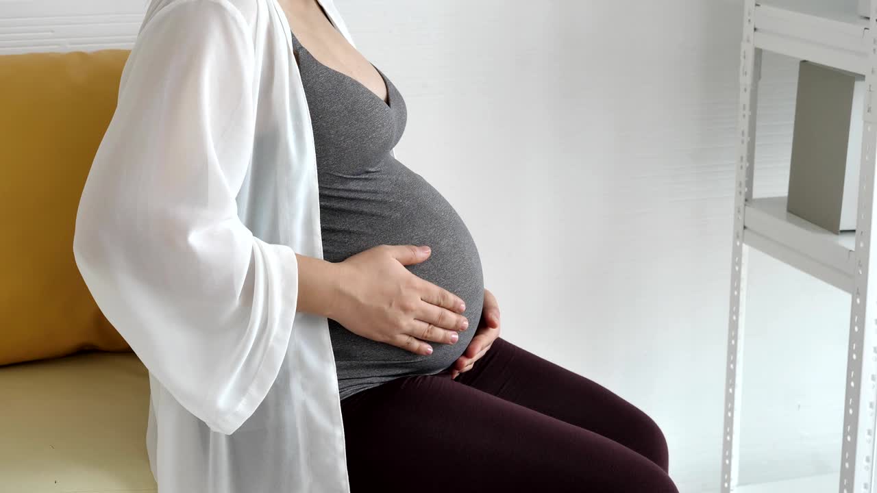 임신한 여성이 집 침실의 창문 근처 소파에 앉아있는 동안 손으로 배를 부드럽게 만지고 있습니다. 사람들이 생활 방식과 건강 관리. 신생아와 산부인과. 어머니와 싱글  개념.