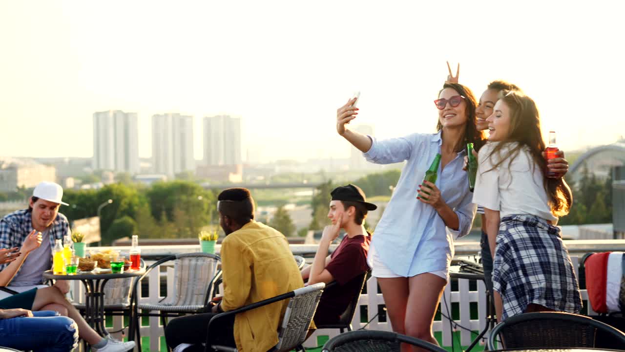 Playful young women are taking selfie with smartphone making funny faces showing tongue and rabbit ears having fun. Girls are wearing trendy clothes and holding bottles.