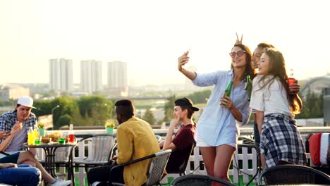 playful young women are taking selfie with smartphone making funny faces showing tongue and rabbit ears having fun. girls are wearing trendy clothes and holding bottles.