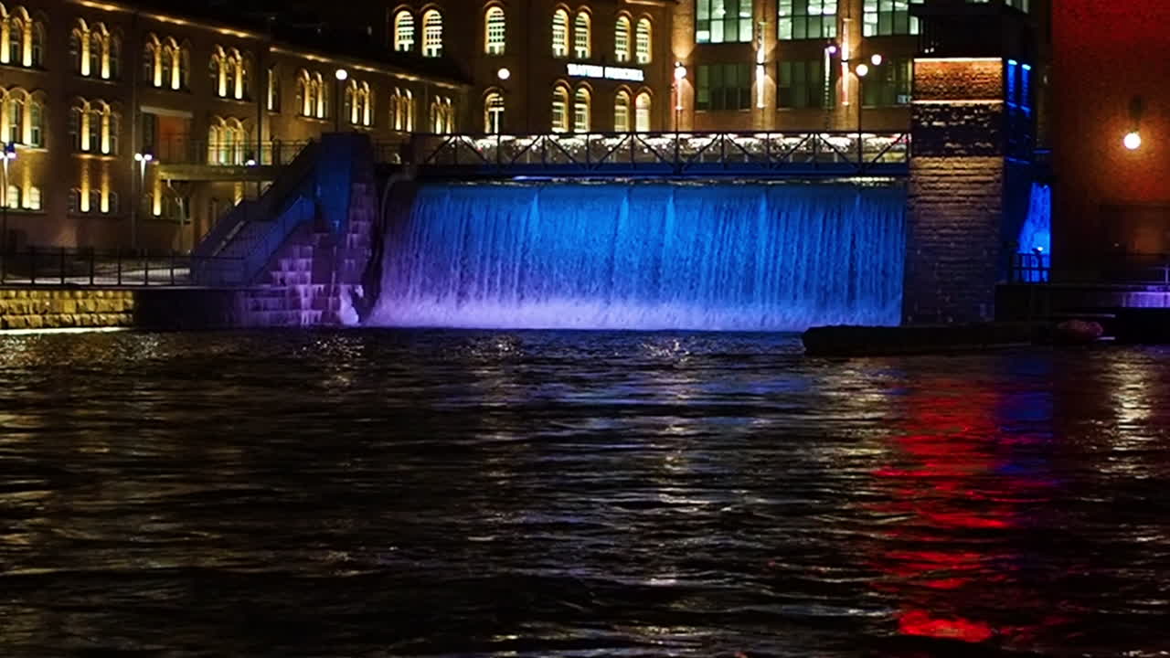 Bridge and waterfall in front of buildings. Water illuminated in blue light.
