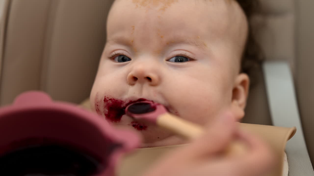 Infant seated in a high chair while an adult feeds the baby with a spoon
