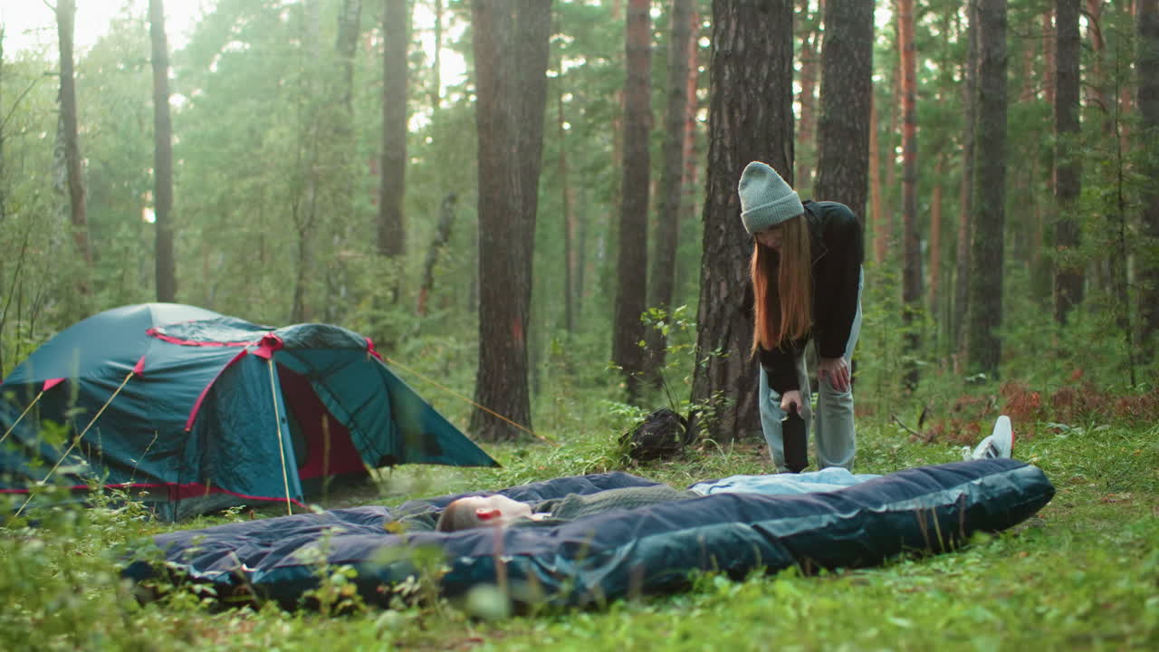 Man lays on tent bag as woman slightly bent pumps air into bag, both looking warmly at each other with soft smiles, tent behind them surrounded by trees