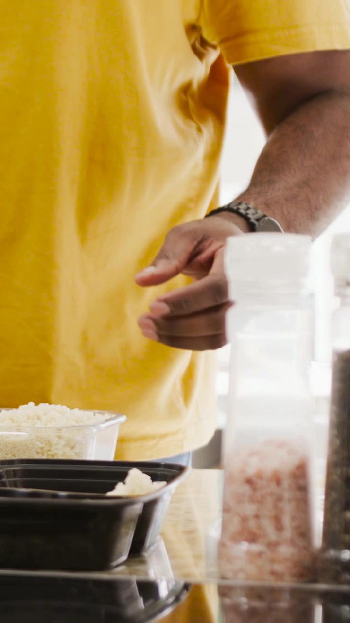 Video verticale: Uomo in maglietta gialla che serve riso, riempiendo contenitori neri per pasti su un bancone con macinini.