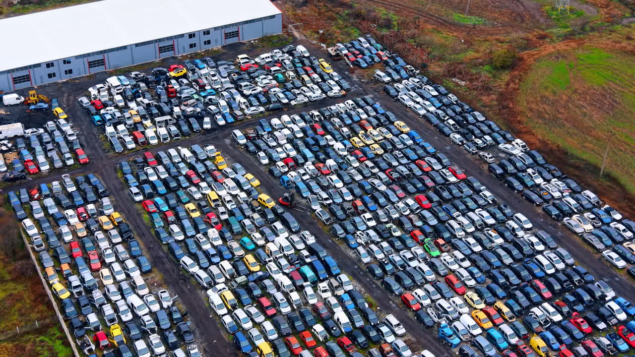Prospettiva aerea diagonale di uno sfasciacarrozze e un magazzino. File di auto incidentate parcheggiate in un impianto di recupero vicino a un grande edificio industriale grigio.