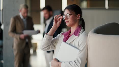 a tired brunette girl in a modern office in a white jacket and pink shirt adjusts her round glasses and looks to the side against of the office and his colleagues