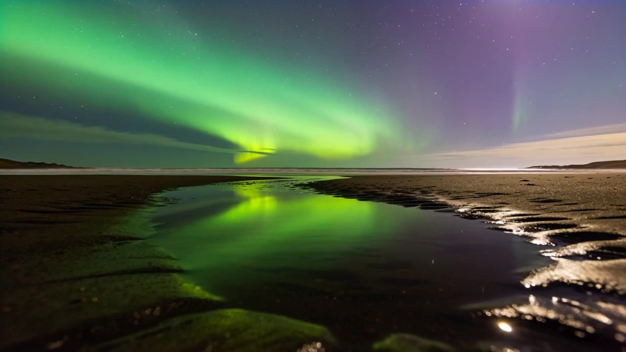 Norrskensridåer framträder och breder ut sig över himlen vid en kuststrand, speglas i en tidvattenpöl.