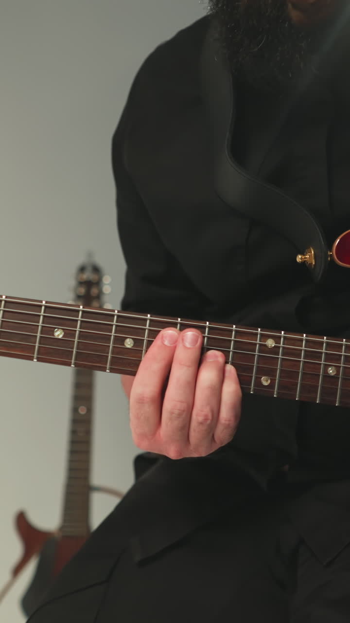un guitarrista con un traje negro, sentado y tocando una guitarra eléctrica roja contra un fondo gris, con otra guitarra visible en el fondo. el músico se centra atentamente en la actuación