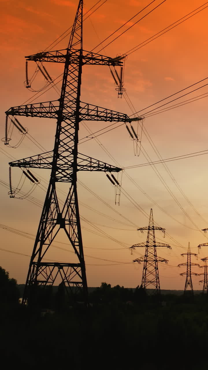Electric towers against orange sky at sunset. High-voltage electricity pylons in the evening. Panoramic view of transmission lines. Camera rising. Vertical video