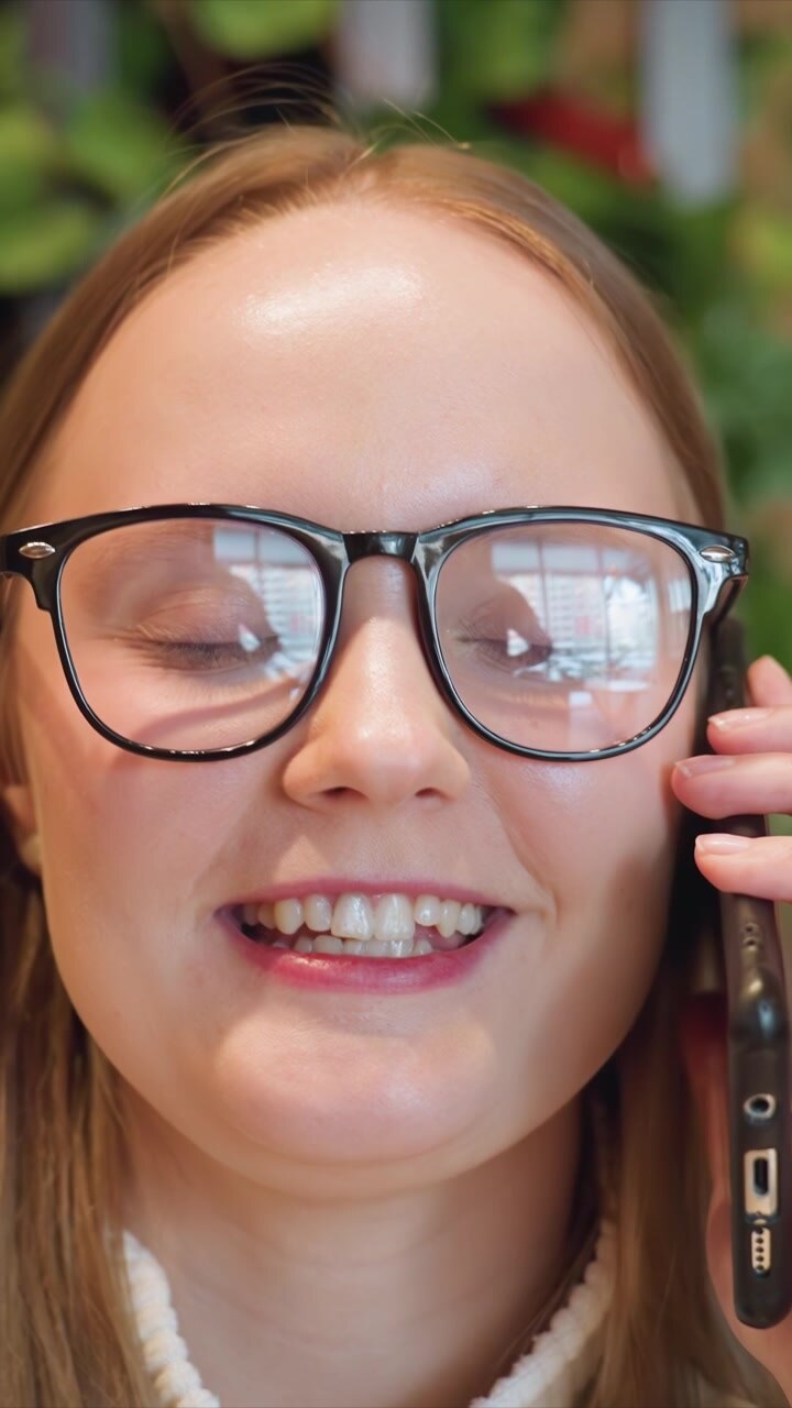 Vista ravvicinata di una donna con gli occhiali che tiene uno smartphone all'orecchio, talvolta insieme a una grande tazza in ceramica. Lo sfondo è costituito da vegetazione leggermente sfocata, che suggerisce un bar al chiuso.