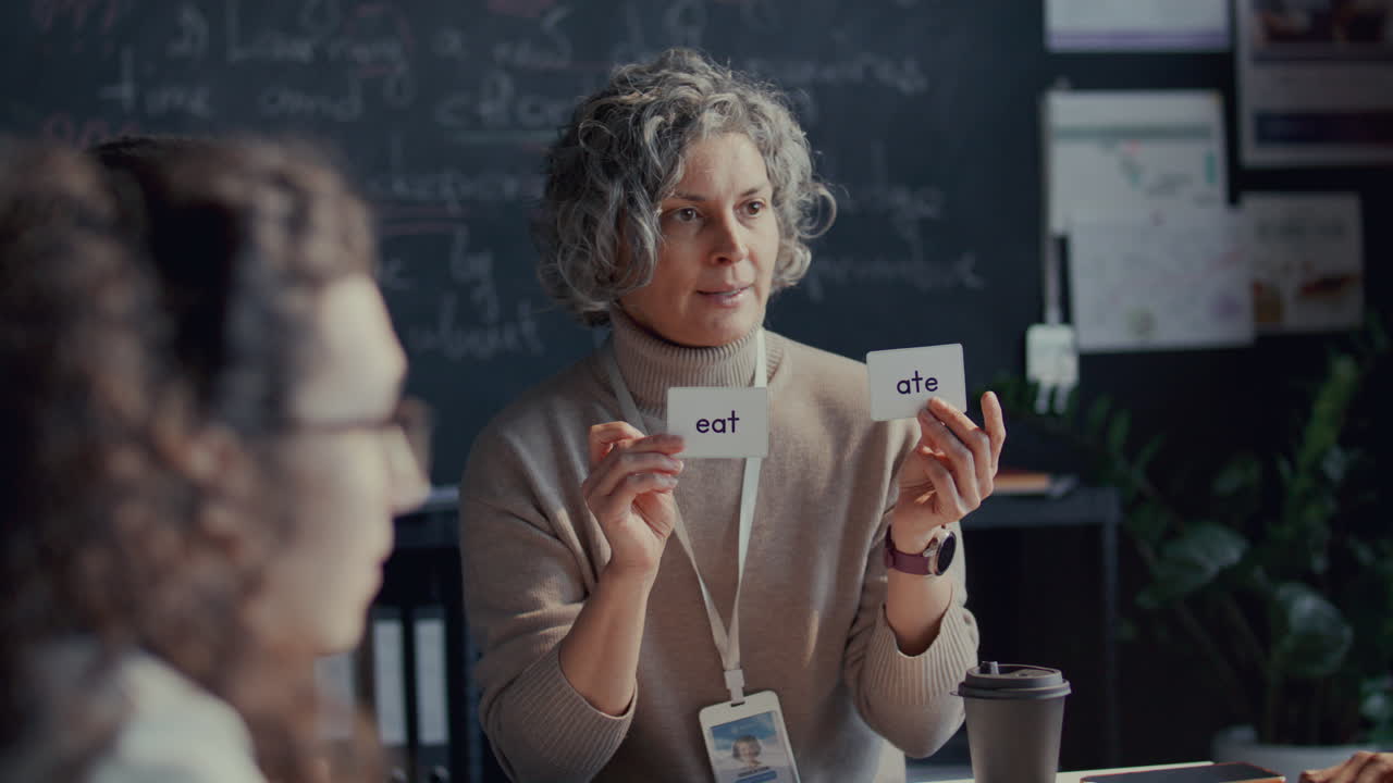 maestra de mediana edad explicando los tiempos verbales con tarjetas de memoria en la clase de inglés