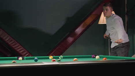 el jugador con camisa blanca y pantalones grises se inclina sobre la mesa de billar verde, agarrando el palo de la clave con un enfoque intenso mientras apunta el tiro. las luces superiores iluminan la sala de billar con una decoración moderna
