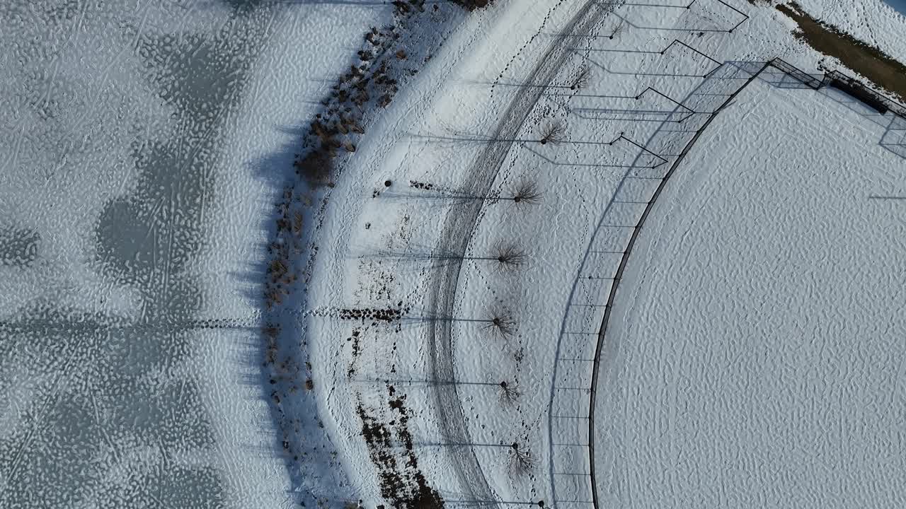 Aerial top-down view of a snow-covered baseball field with trees along the perimeter in winter. The ground is blanketed in snow, and the field's shape is clearly visible.