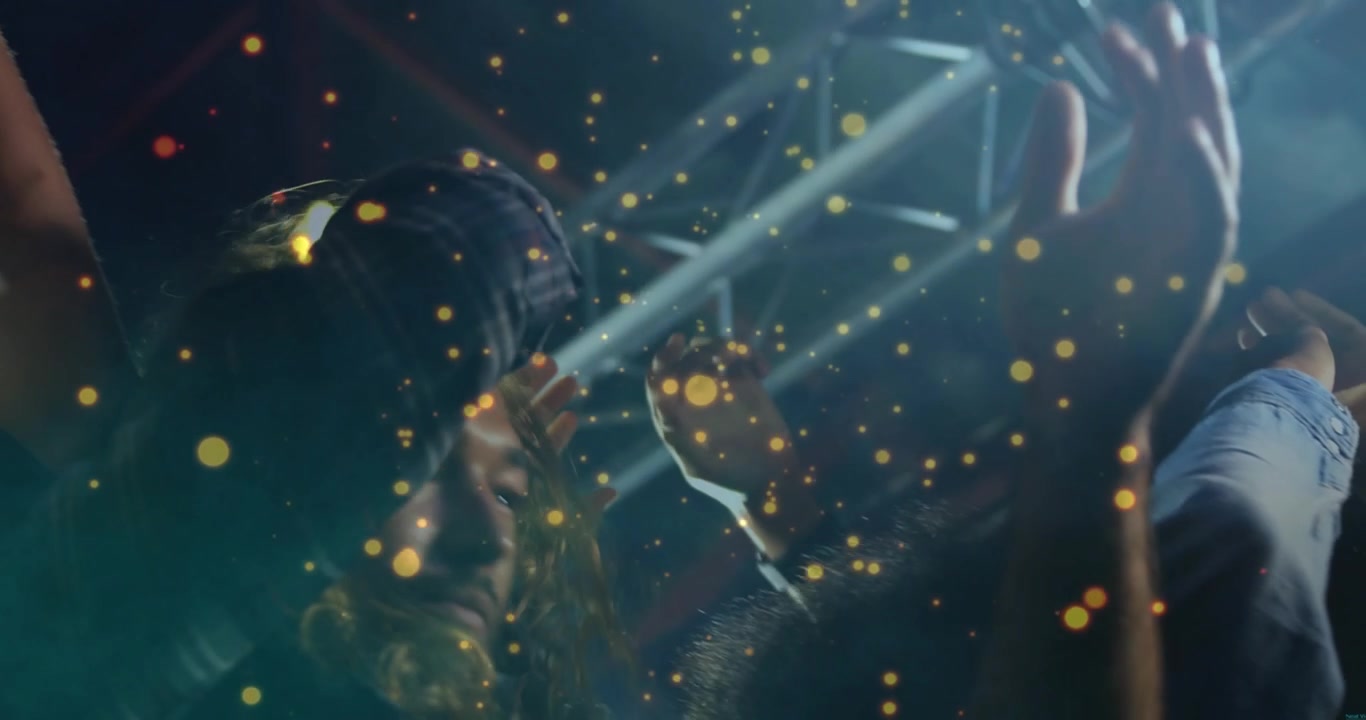 Crowd raising hands in concert, gold particles drifting as sweeping stage lights revealing faces