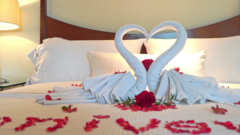 hotel bedroom with fresh towels used as a heart symbol or two swans, word "anniversary" inlaid with red rose petals on the white sheets