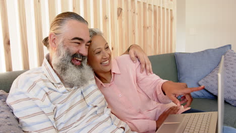 senior couple sitting on couch, smiling and embracing while using laptop
