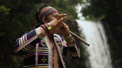 orbit shot of indigenous person playing kubing tribal instrument, behind is a waterfall while standing