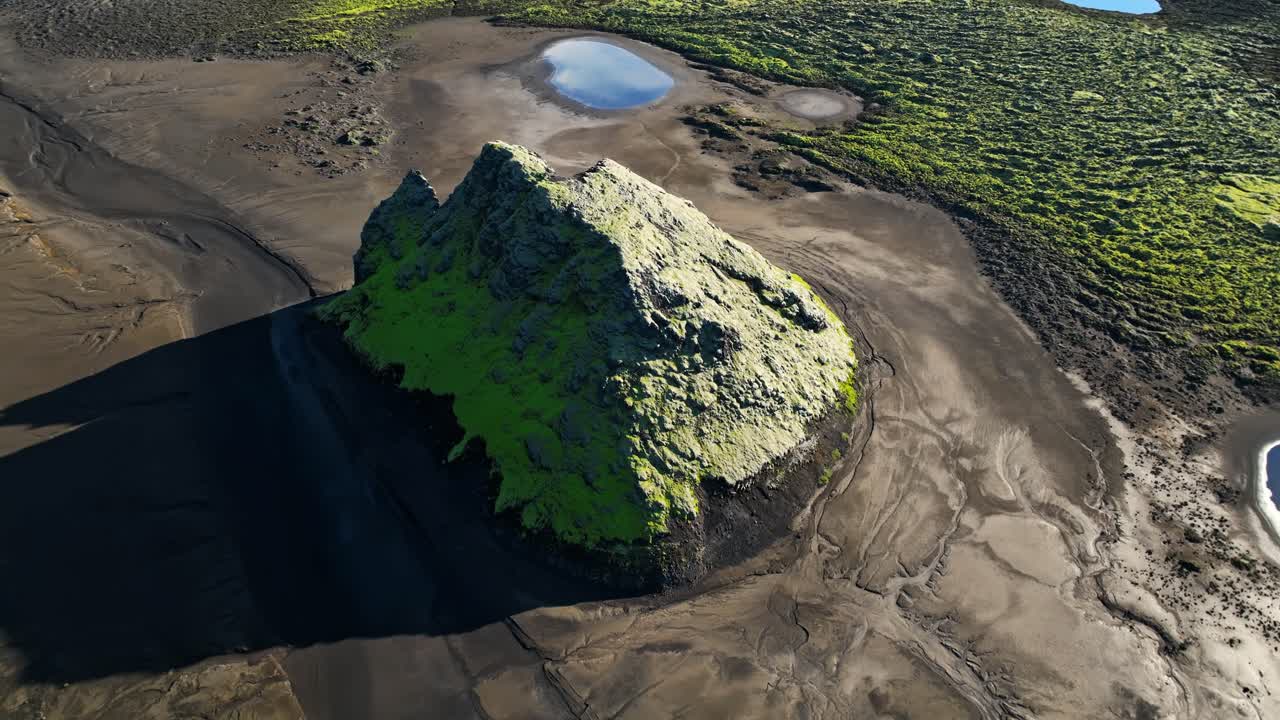 Tiefer Drohnenflug über einen moosbedeckten Vulkanhügel, der sich aus schwarzen Sandebenen im isländischen Hochland erhebt, mit kleinen Wasserbecken und dramatischem Schatten, episches abgelegenes Gelände