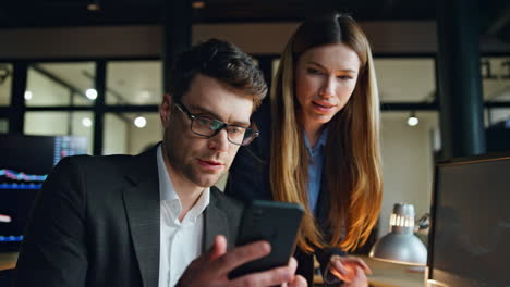 Two-business-people-working-dark-finance-office.-Focused-man-using-smartphone