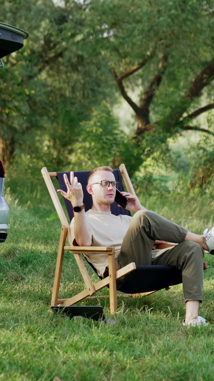 Relaxed male speaks on the phone sitting in the folding chair. Remote work conversation in the woods. Vertical video