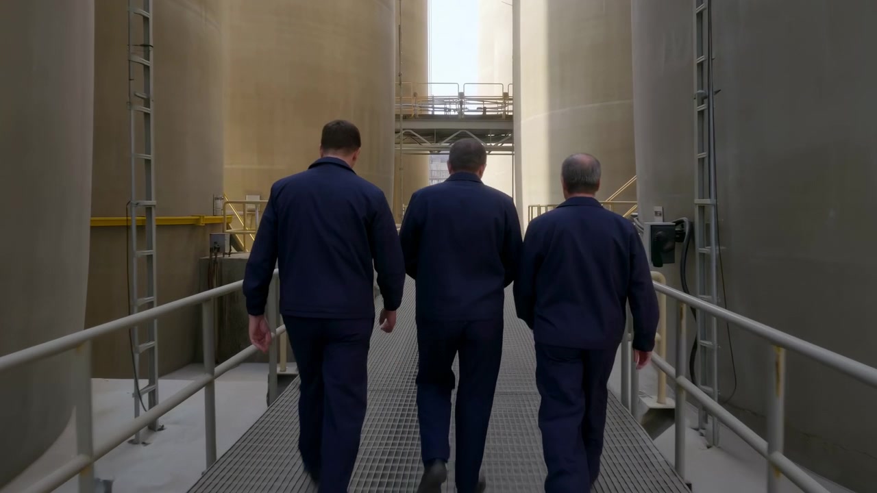 Vista posteriore di tre operai industriali con tute da lavoro blu scuro abbinate che camminano lungo una passerella metallica grigliata tra imponenti serbatoi di stoccaggio cilindrici beige e grigi.