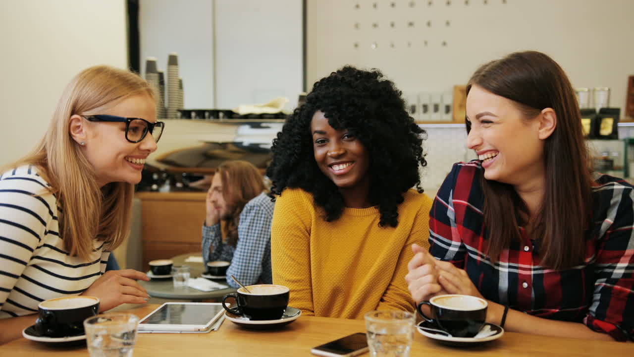 amigas afroamericanas y caucásicas chismeando y riendo sentadas en una mesa mientras usan una tableta en un café
