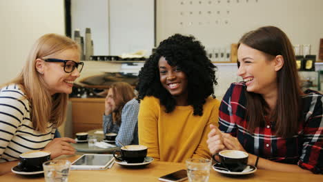 african american and caucasian women friends gossiping and laughing sitting at a table while using a tablet in a cafe