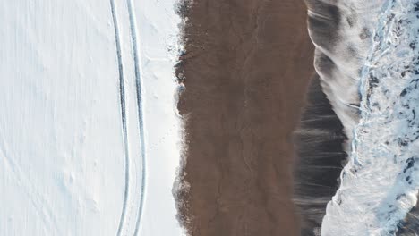 waves break on shore of snow covered sandvik beach in iceland, top down