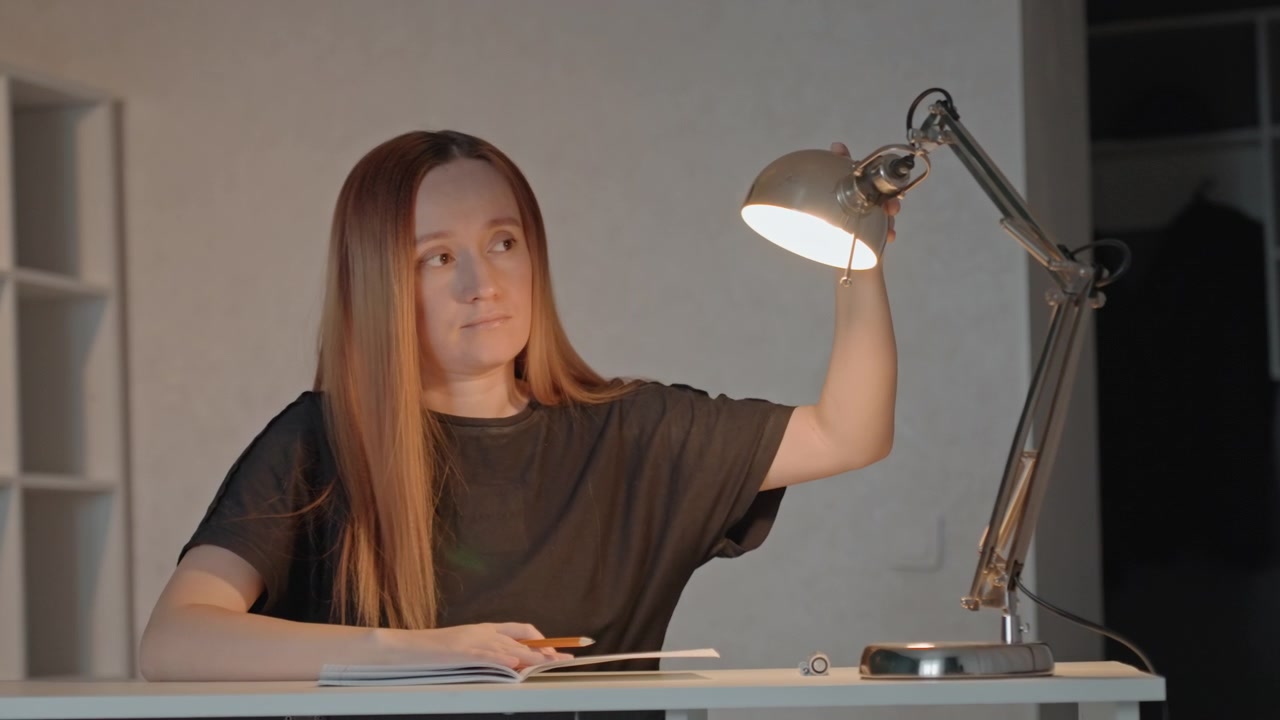 Una giovane donna con i capelli lunghi siede alla scrivania tenendo degli oggetti. La donna interagisce con un libro aperto e una matita in una stanza dai colori neutri. L'illuminazione è costante mentre la donna si concentra sul suo lavoro.