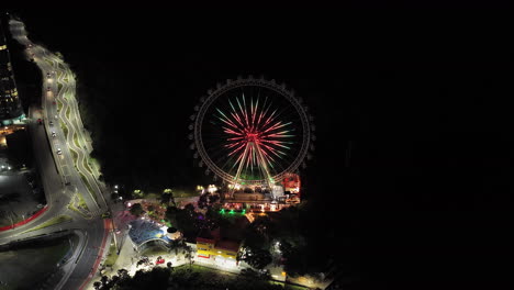 Aerial night view of the brightly lit Ferris wheel in Barra Sul, Balneário Camboriú, Brazil, with surrounding roads, buildings, and festive park lights creating a vibrant nighttime atmosphere