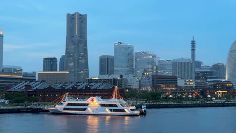 Stadtbild-In-Der-Dämmerung-Mit-Einer-Beleuchteten-Yacht-Am-Wasser,-Wolkenkratzern-Und-Ruhigem-Wasser,-Heitere-Stadtszene