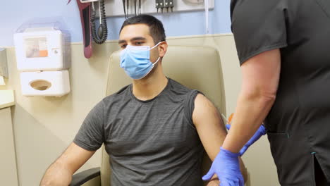 nurse cleans shoulder and administers covid 19 vaccine shot in the arm of young adult dark skinned man of minority ethnicity wearing a medical mask, corona virus injection in doctor’s office