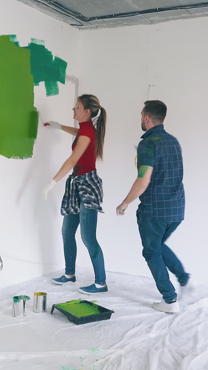 Dolly cam shot: positive couple in dirty clothes has fun walking around tray and painting white wall in spacious renewing room