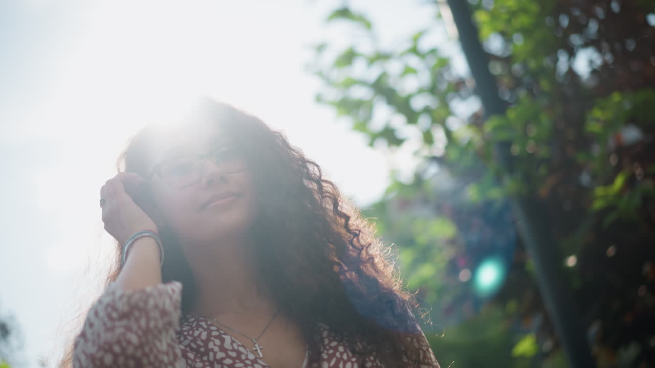 mujer con gafas y colgante de cruz posa al aire libre, sonriendo cálidamente y ajustando su cabello mientras la luz del sol se refleja en su cara, rodeada de exuberante vegetación y suave luz natural