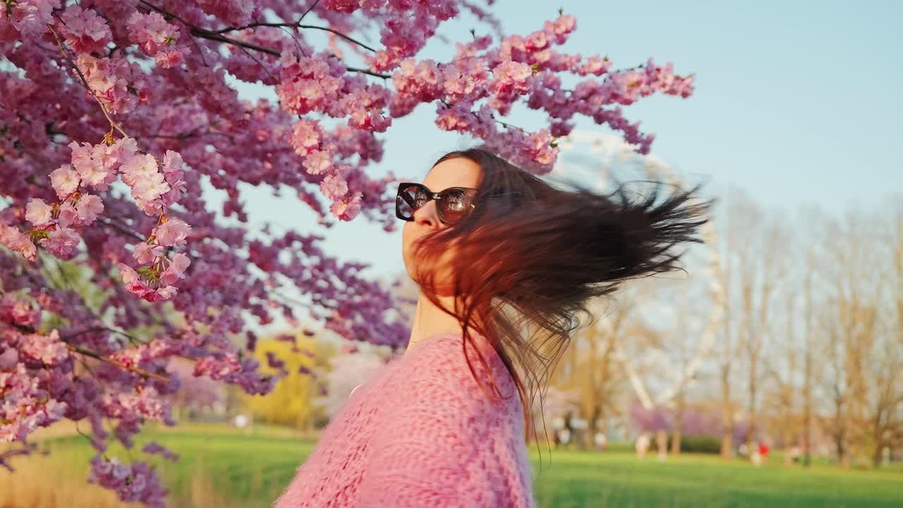 Slow motion turn with flowing hair beneath cherry trees near ferris wheel, dusk