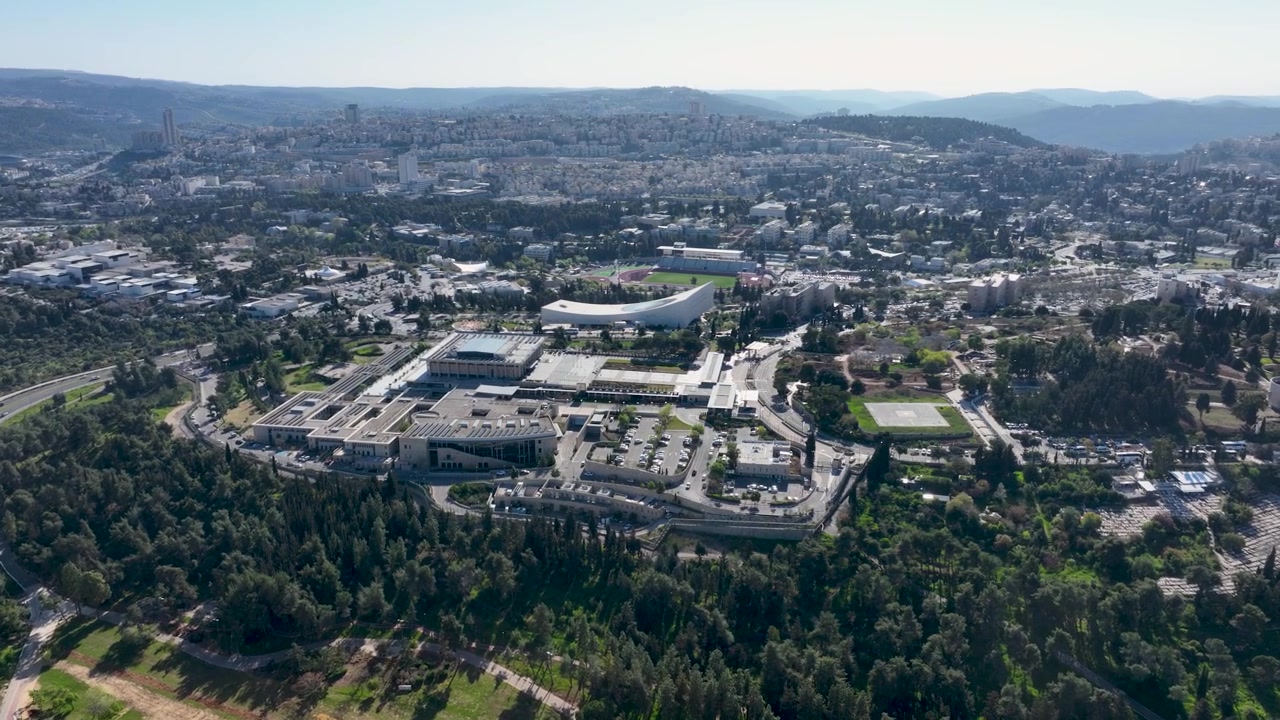 Ampia vista aerea del campus Givat Ram dell'Università Ebraica circondato da una fitta vegetazione forestale e dal paesaggio urbano di Gerusalemme sotto la chiara luce del giorno