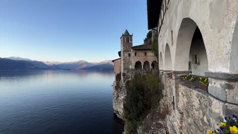 panning view of santa caterina del sasso hermitage in italy perched on lake maggiore with snowy alps mountains range in background