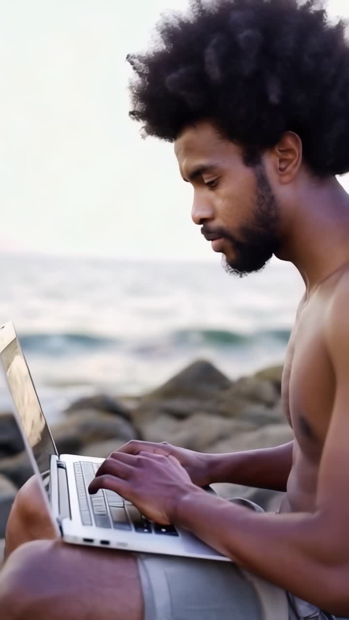 A man is sitting on a rock by the ocean, typing on a laptop