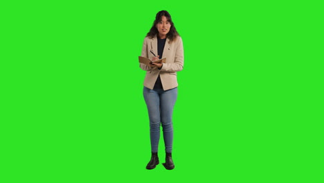 full length studio portrait of female teacher with notebook taking class register standing against green screen