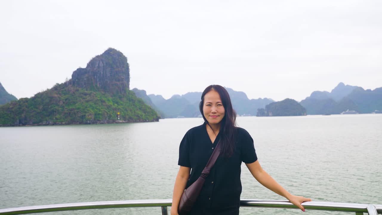 Asian woman stands at the bow of a cruise ship, gazing contemplatively at the dramatic limestone karsts and emerald waters of Ha Long Bay, Vietnam, - front view looking at camera