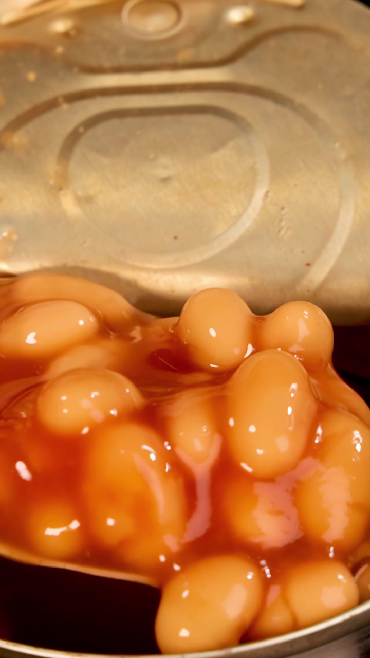 Uma colher retira feijão cozido em molho de tomate de uma lata de metal aberta sob iluminação quente e uniforme. A perspectiva em close-up destaca a textura e a superfície brilhante.