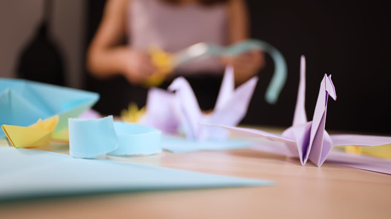A person uses yellow scissors to cut paper strips at a desk with origami cranes