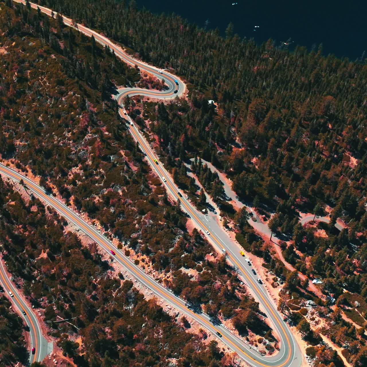 Twisted road through the wooded rocky landscape with many cars moving by. Deep blue lake at the foot of mountain from aerial perspective