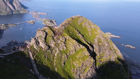 Reinebringen-peak-aerial-view,-Travelers-walking-down-the-hill-following-wavy-mountain-trails,-distant-view-of-Reine-villages-and-Lofoten-Islands