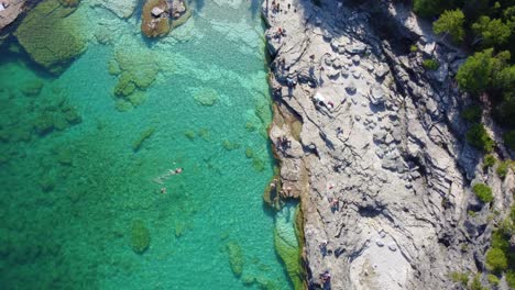 Overhead-drone-shot-tilting-slowly-to-reveal-the-rest-of-the-scenery-of-a-rocky-coastline-with-people-swimming-off-the-coast-of-Georgian-Bay,-in-Ontario,-Canada